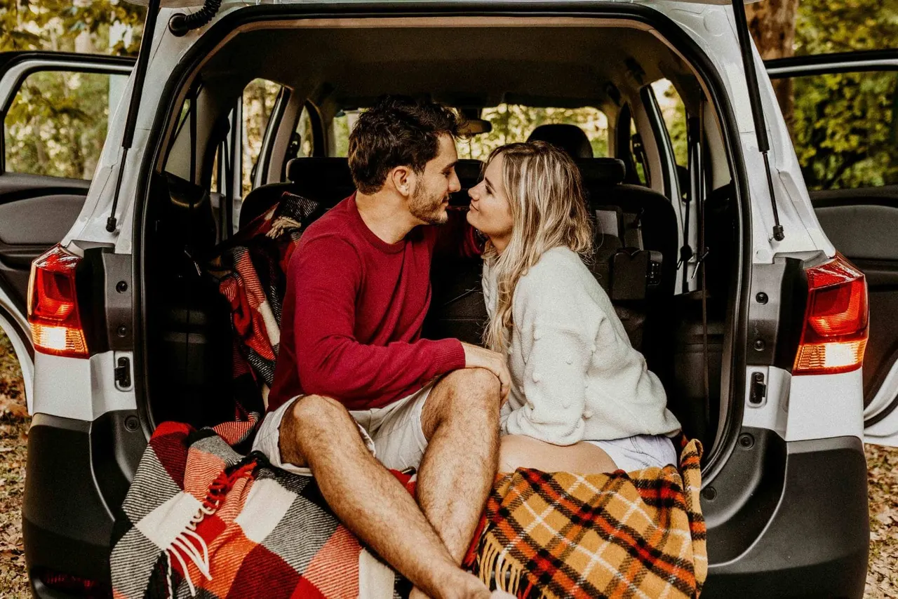 A couple sitting close in the open boot of a car wrapped in blankets