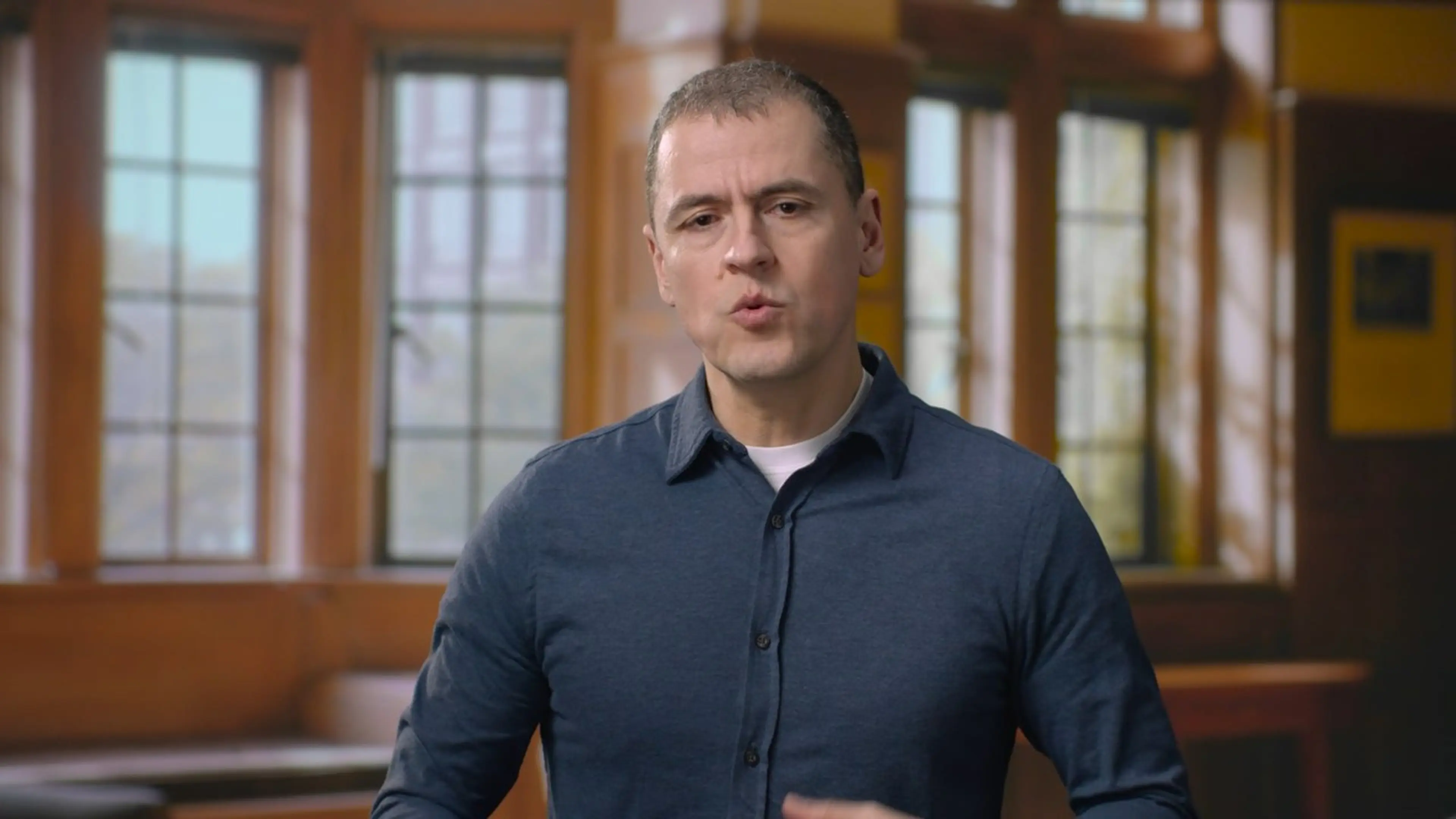 Live-action video production still — a speaker filmed on location in a wood-paneled office