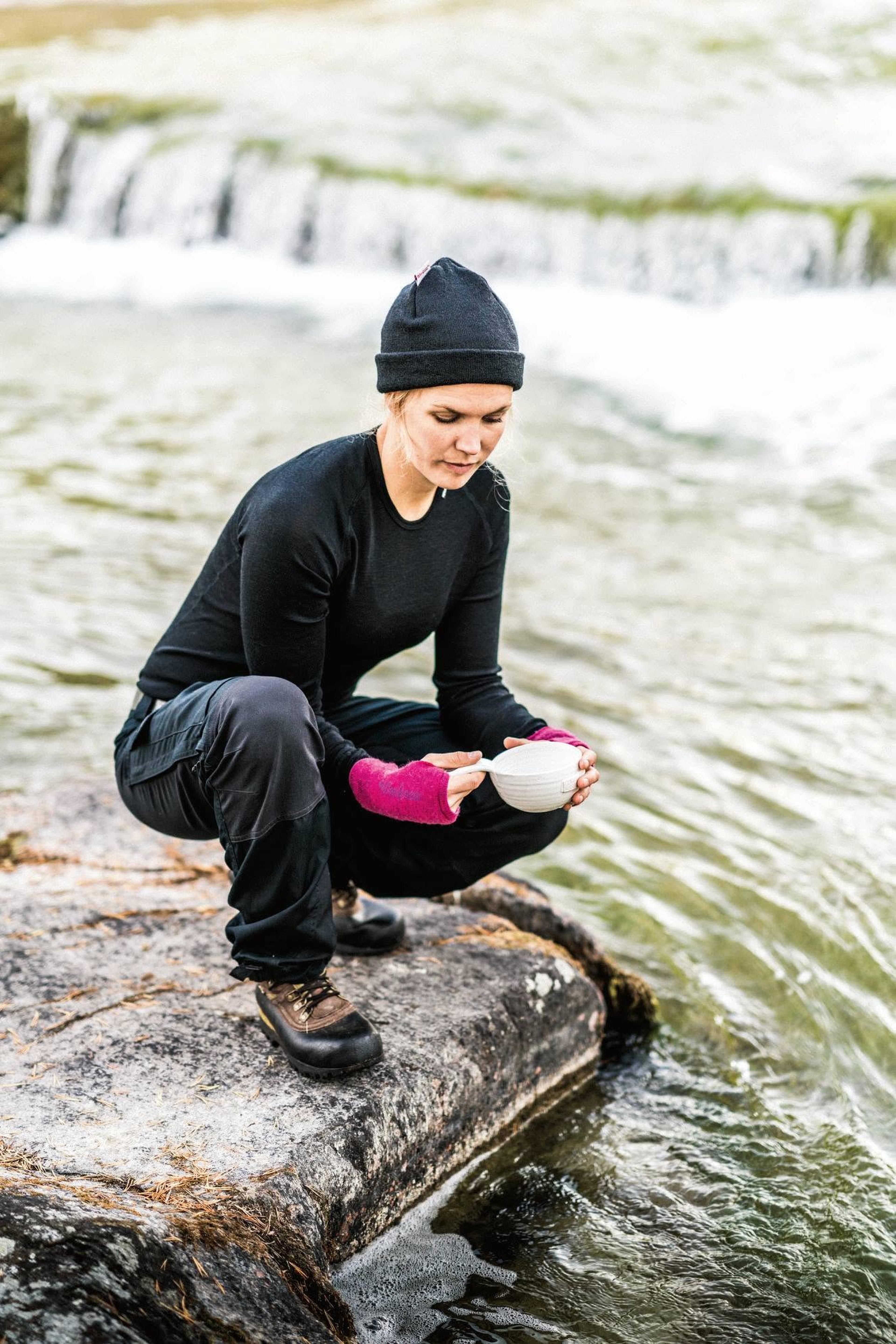 A person in outdoor attire crouches on a rock by a river, holding a white bowl, with a waterfall visible in the background.