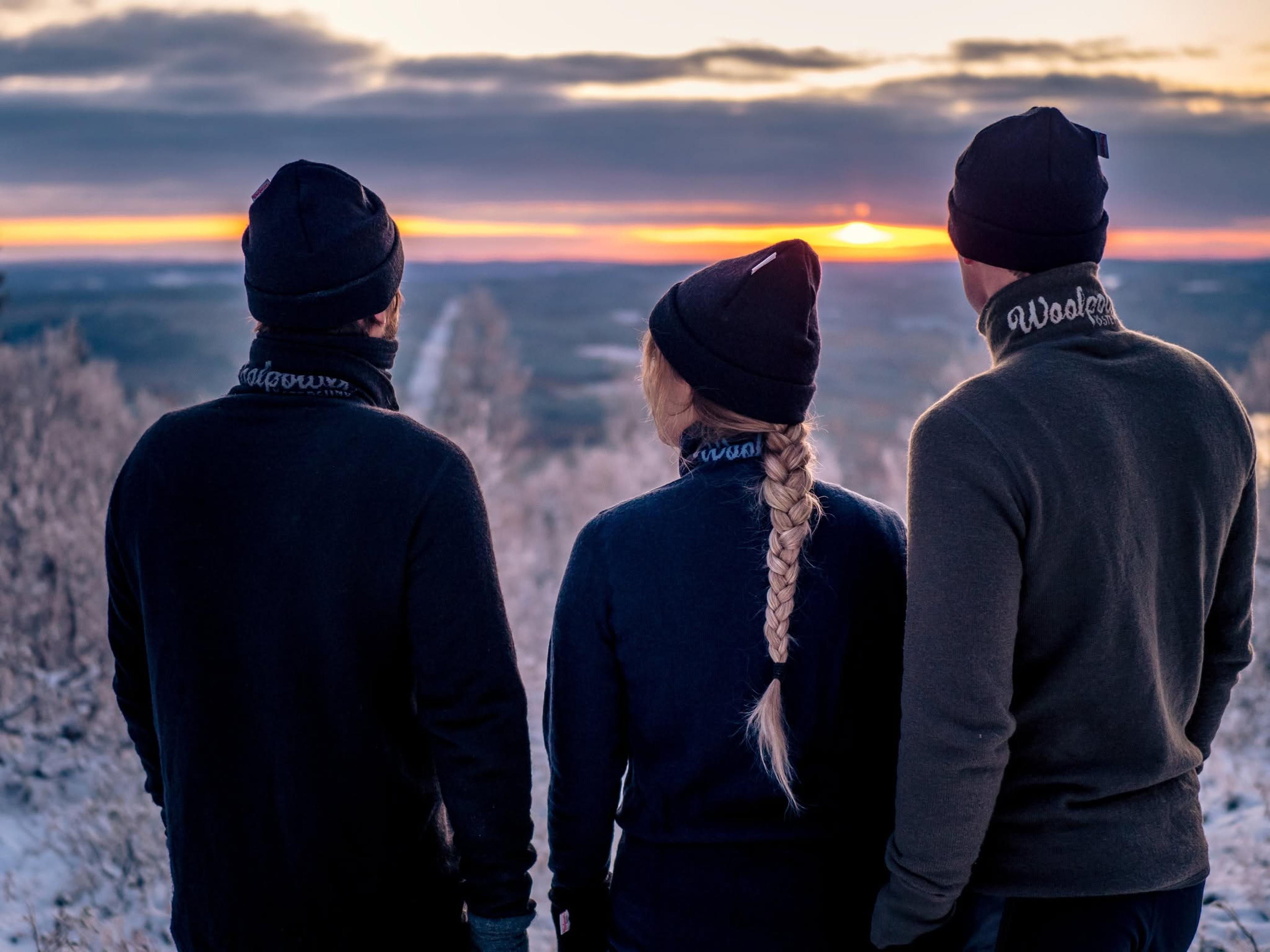 Three people in winter clothing stand on a snowy hill, facing a vivid sunset over a vast landscape.