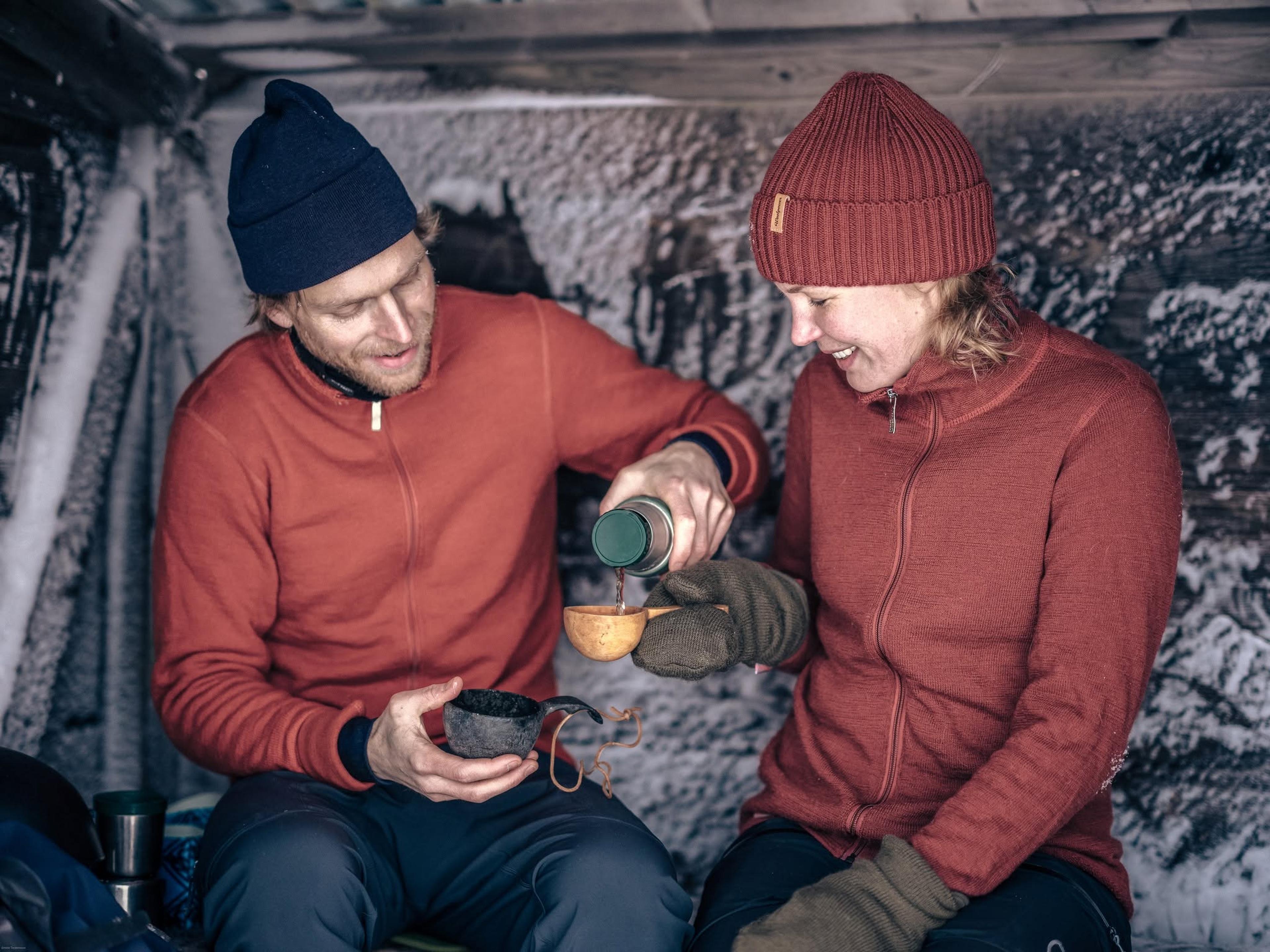 Two people in matching red jackets and hats pour a hot drink in a snowy shelter, smiling warmly at each other.