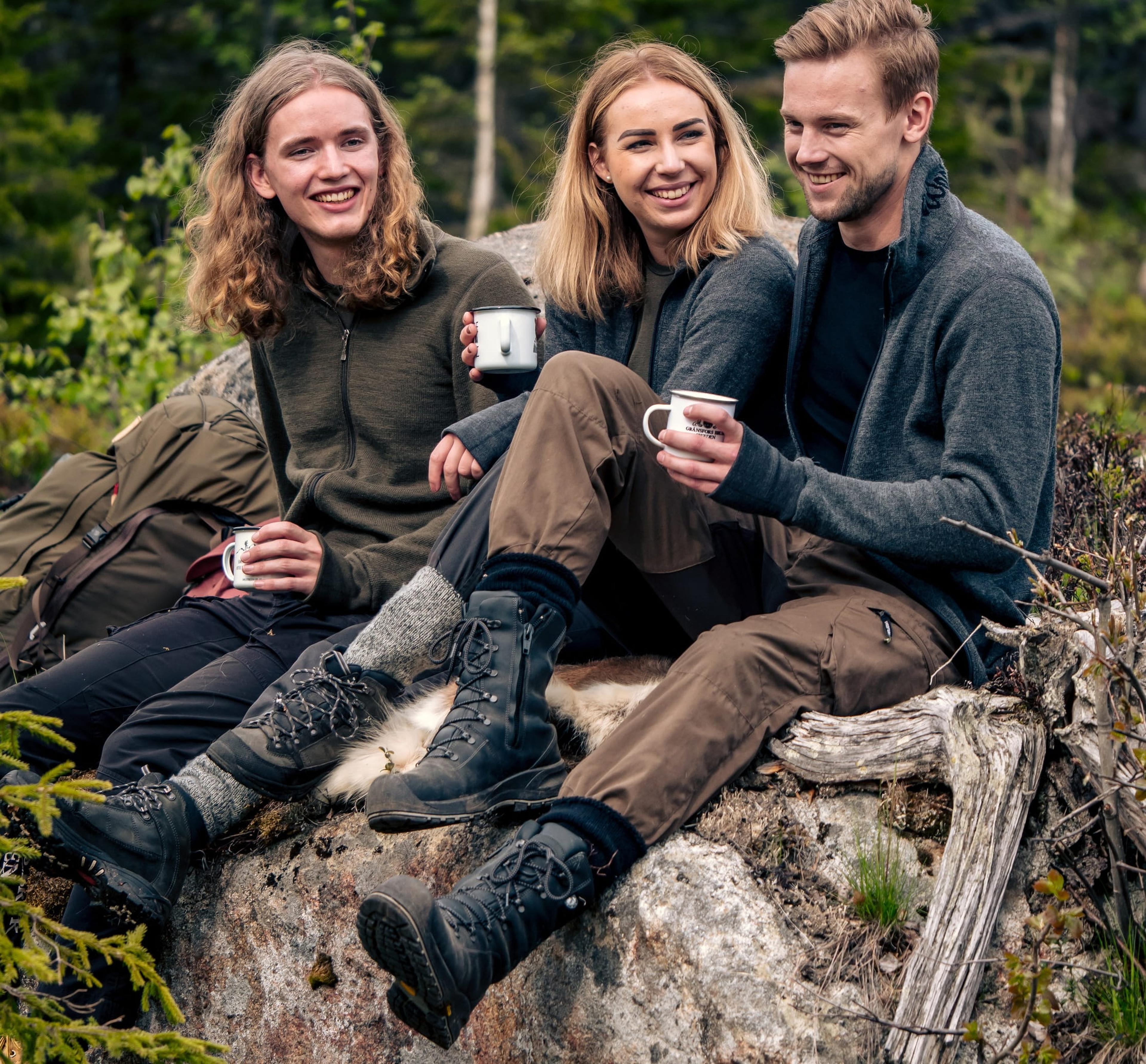 Three people sitting on a rock in the forest, smiling and holding mugs, wearing outdoor clothing and hiking boots, with backpacks nearby.