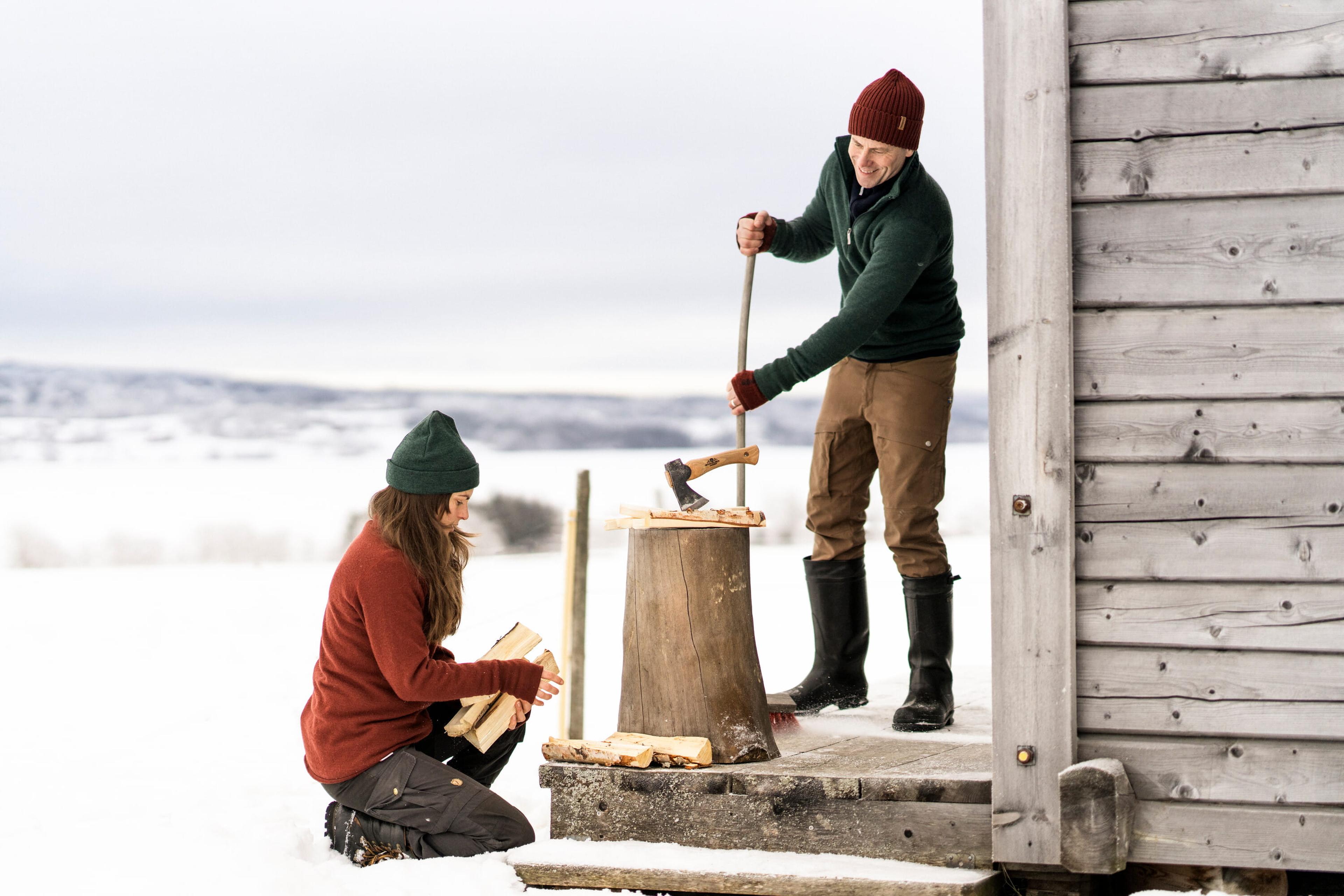 Two people chopping wood on a snowy day, beside a wooden cabin. One is kneeling with logs, the other is using an axe on a stump.