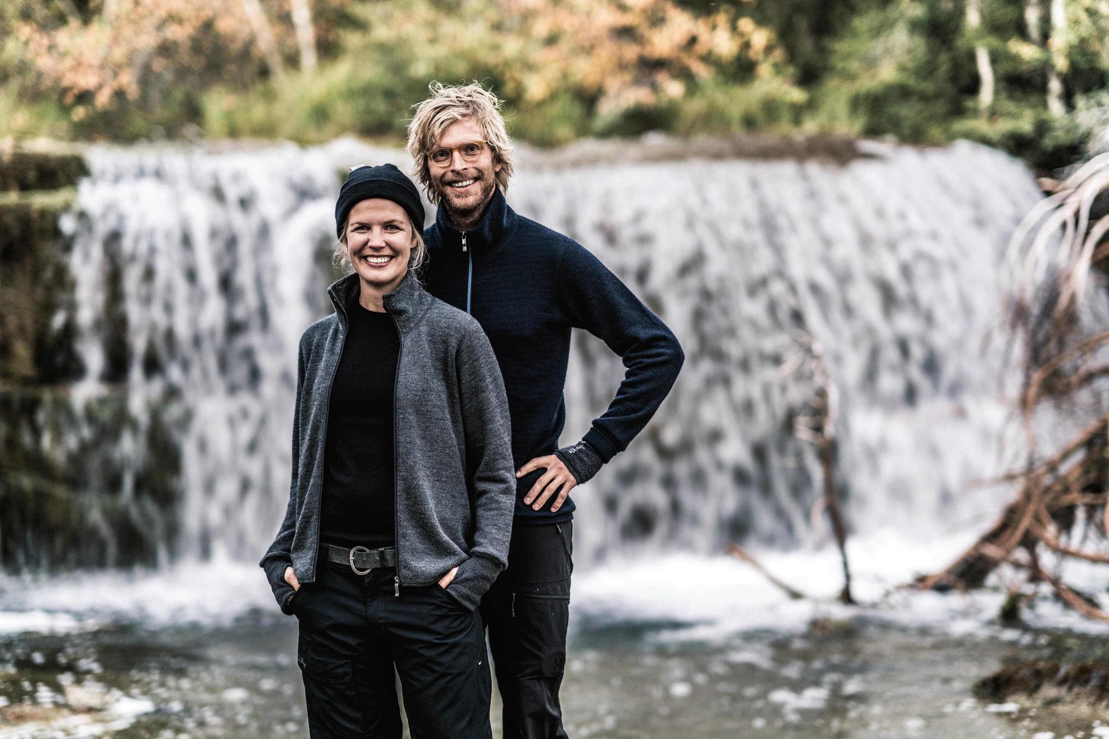 Two smiling people in outdoor attire standing in front of a waterfall, surrounded by greenery.