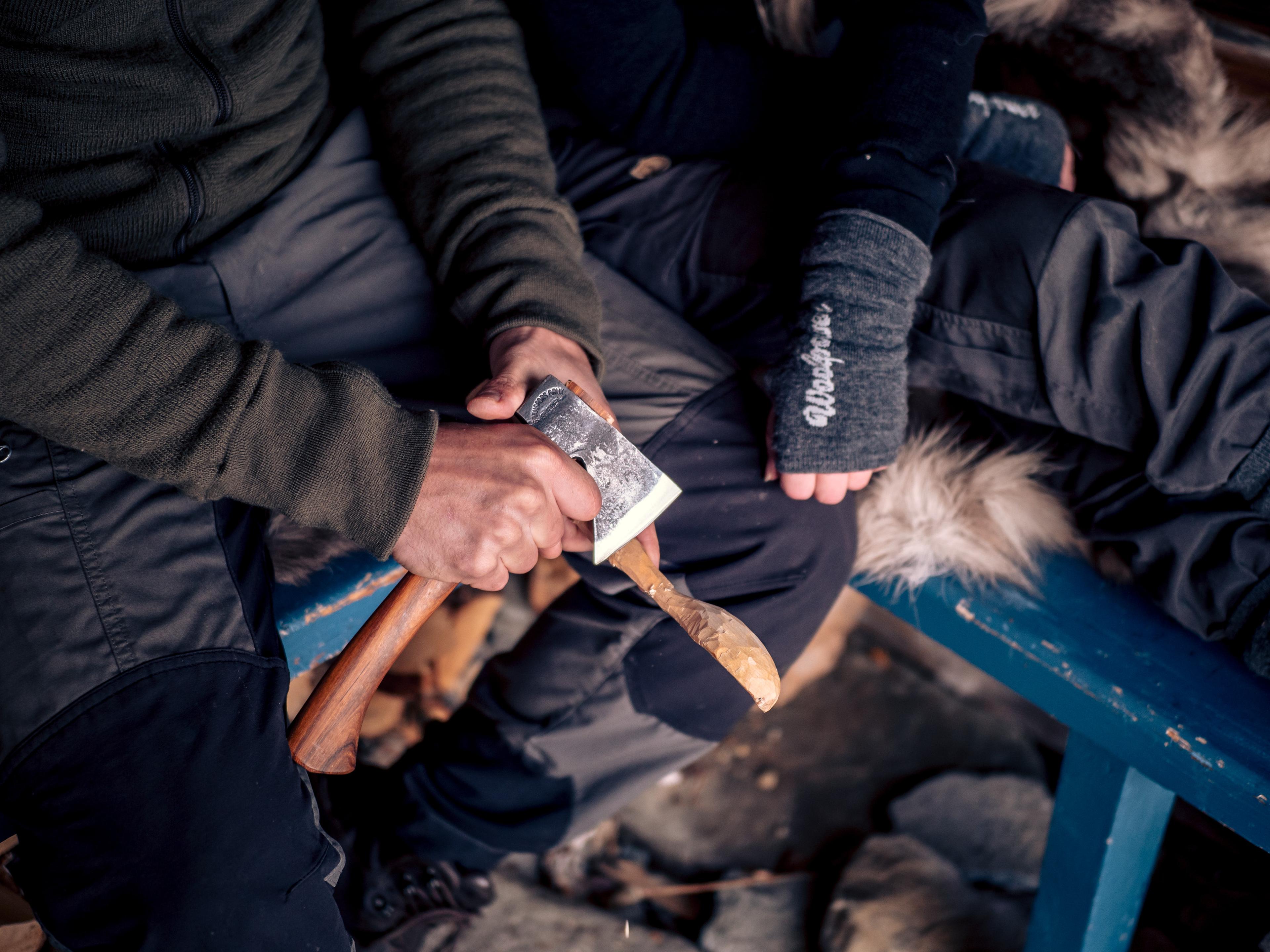 Two people sitting on a bench carving wood with an axe while wearing Grey Gaiters