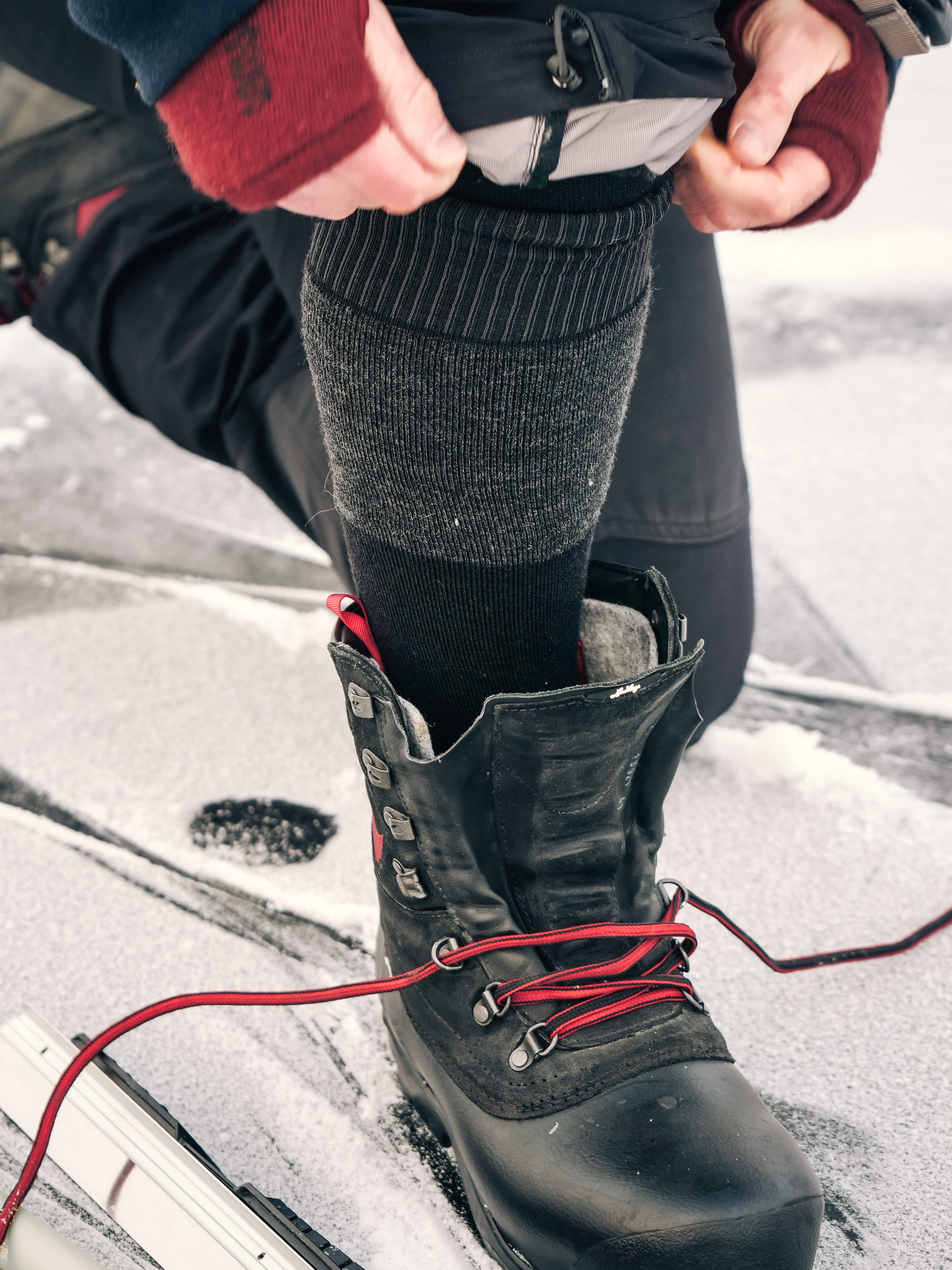 Person adjusting gray sock and black snow boot with red laces on a snowy surface.