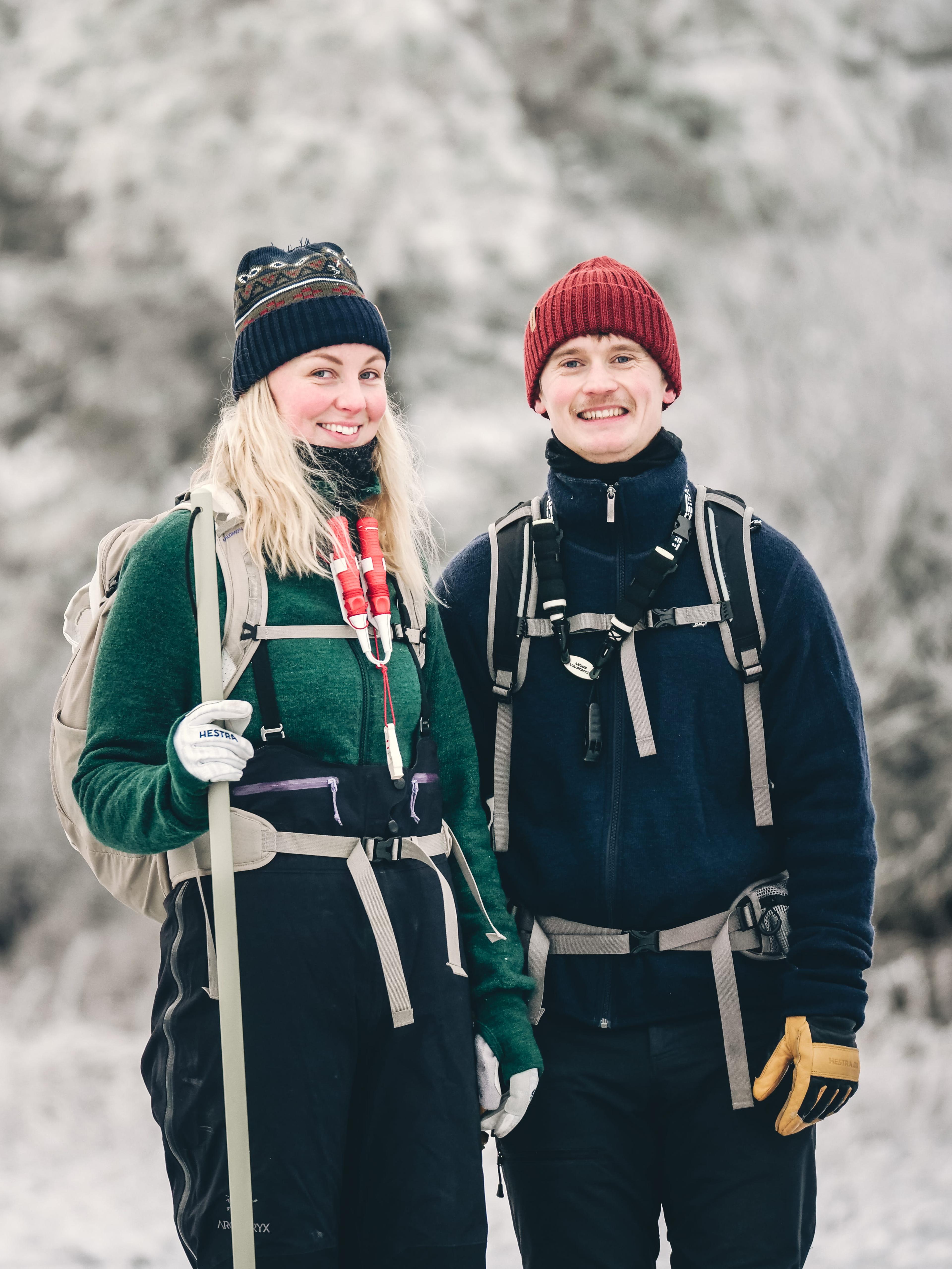 A man and woman in winter clothing, smiling outdoors while hiking in a snowy landscape, each wearing backpacks and knitted hats.