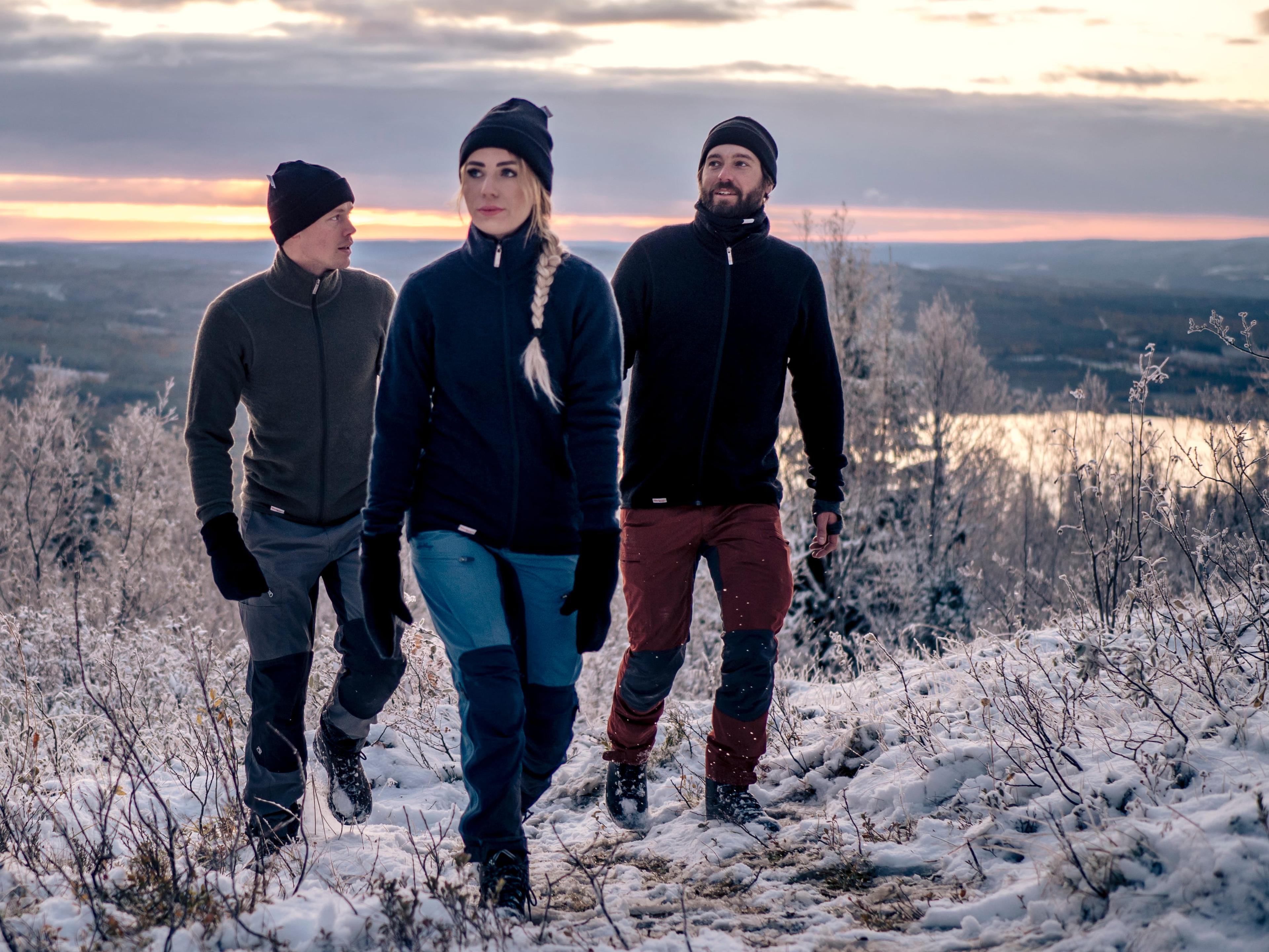 Three people wearing winter clothing hike through a snowy landscape at sunset, with a frozen lake and distant hills in the background.