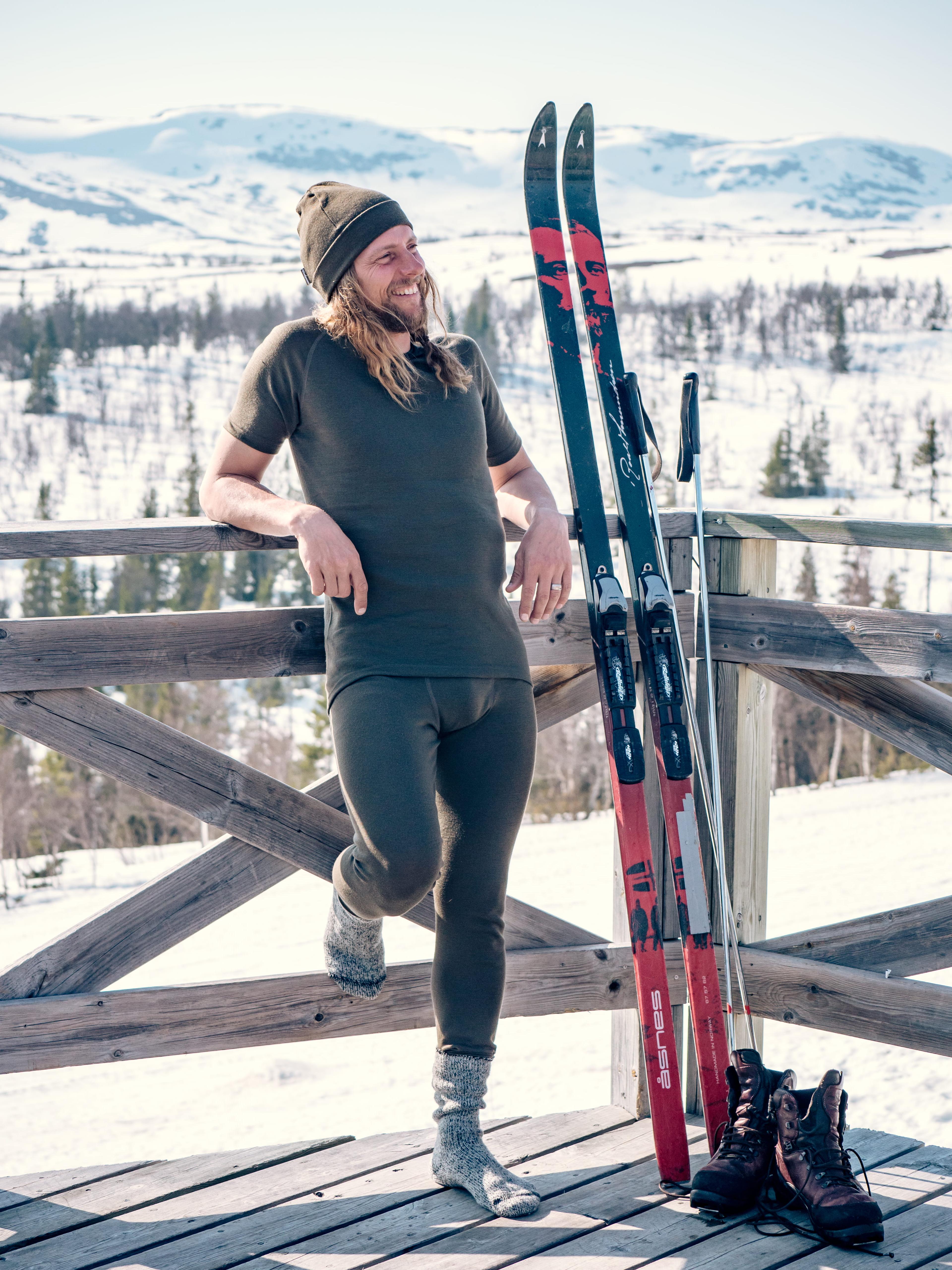 A man in thermal wear and a beanie leans on a wooden fence, smiling in snowy mountains, with skis and boots beside him.