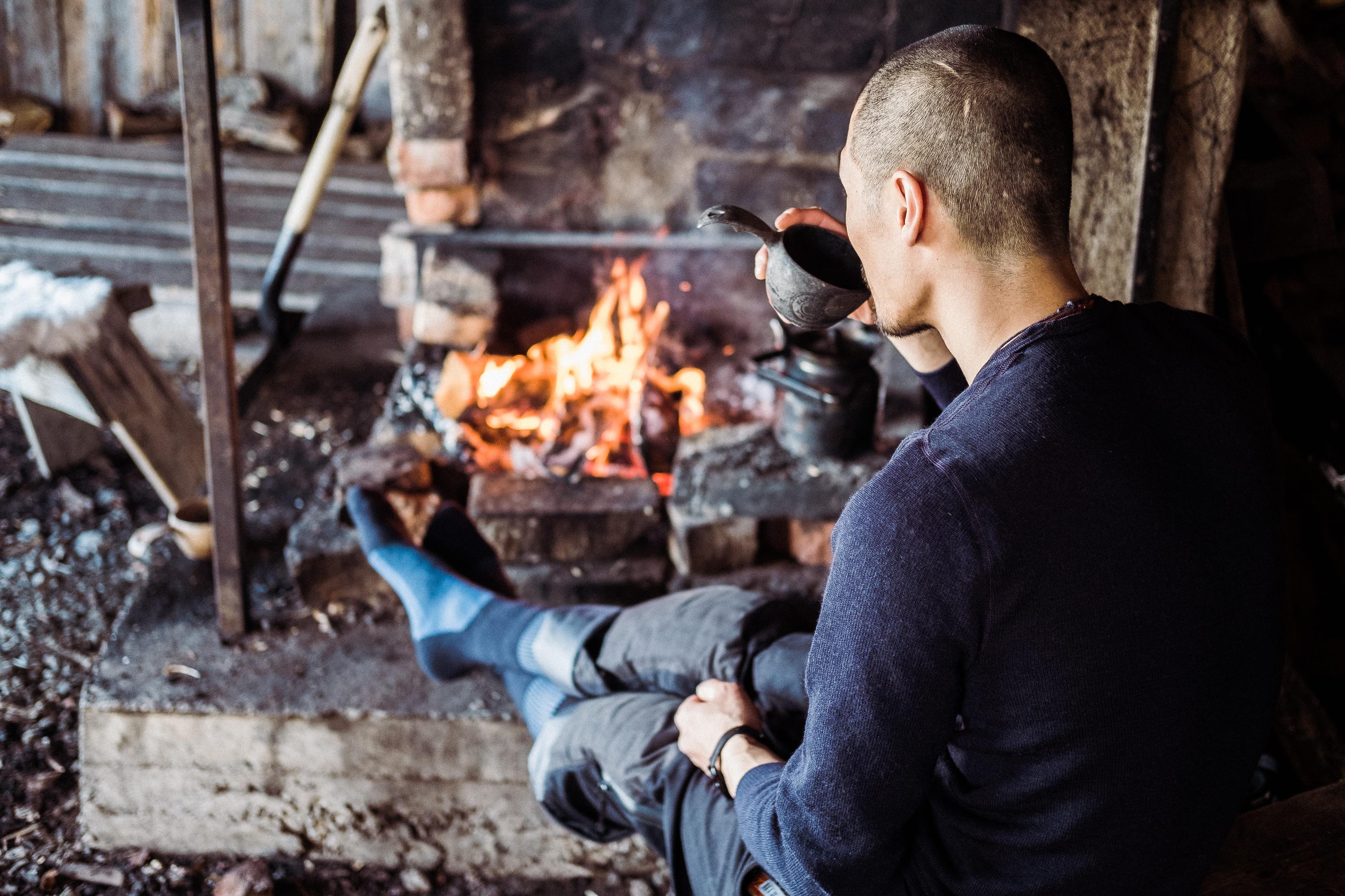 A person with short hair sits by a cozy fireplace, wearing blue socks, drinking from a cup, and enjoying the warmth.