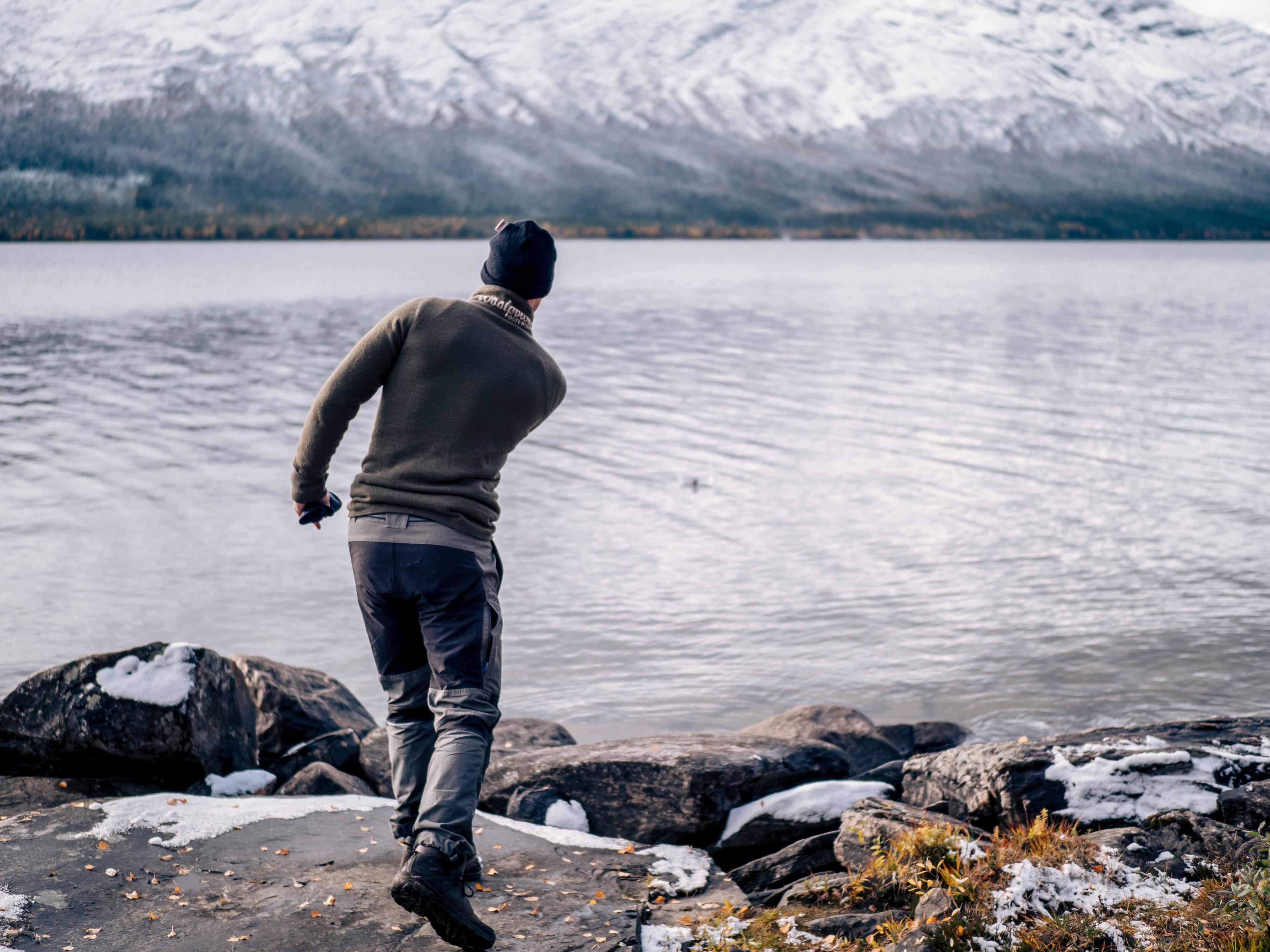 A person in a sweater and cap skips stones on a calm, snow-lined lake with mountain views.