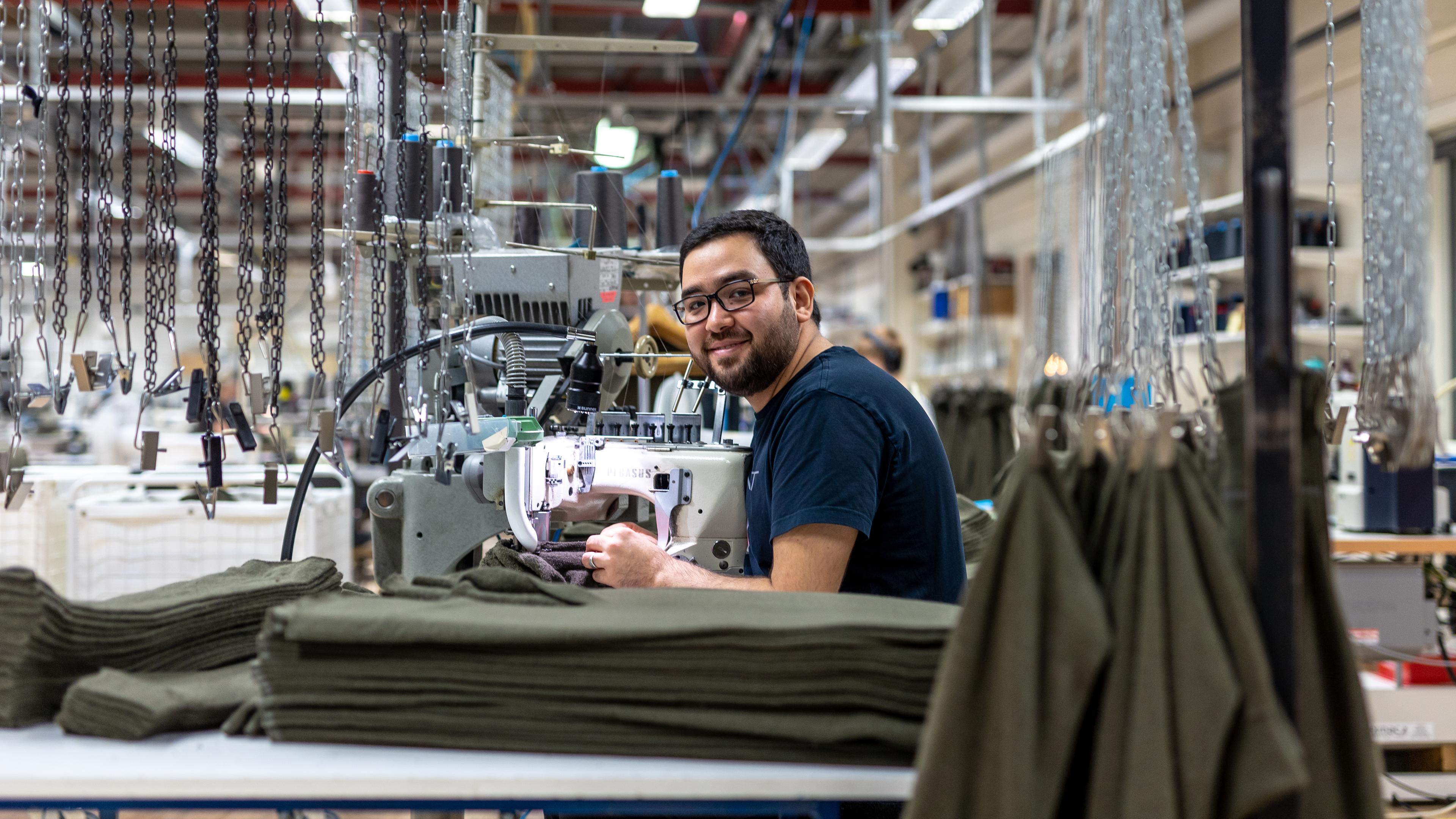 Man operating a sewing machine in a textile factory, surrounded by fabric stacks and industrial equipment.