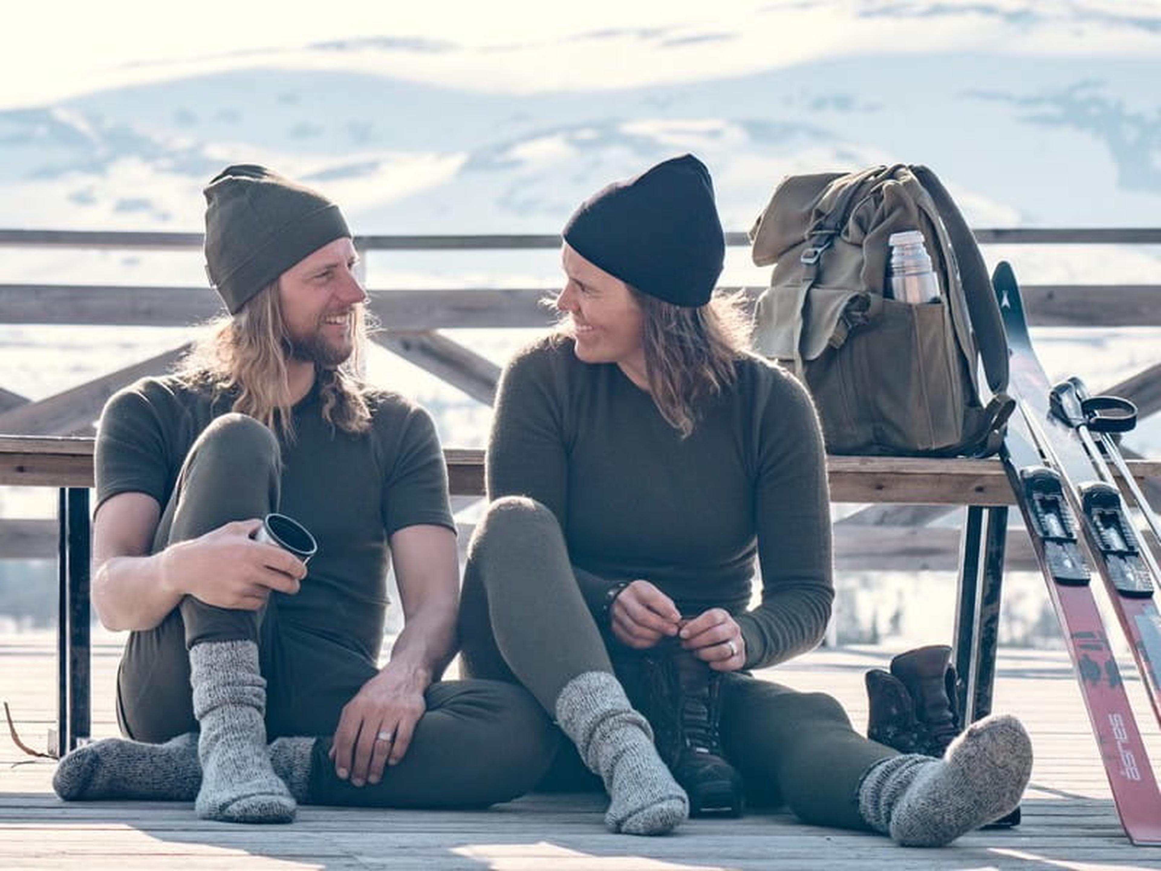 A man and woman sit on a bench, both holding skis, with a snowy landscape in the background.