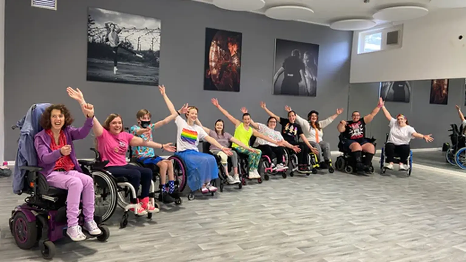 A group of people in wheelchairs joyfully raise their arms in a dance studio, surrounded by wall art and mirrors.