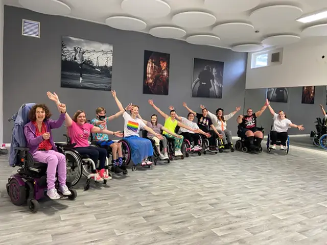 A group of people in wheelchairs joyfully raise their arms in a dance studio, surrounded by wall art and mirrors.
