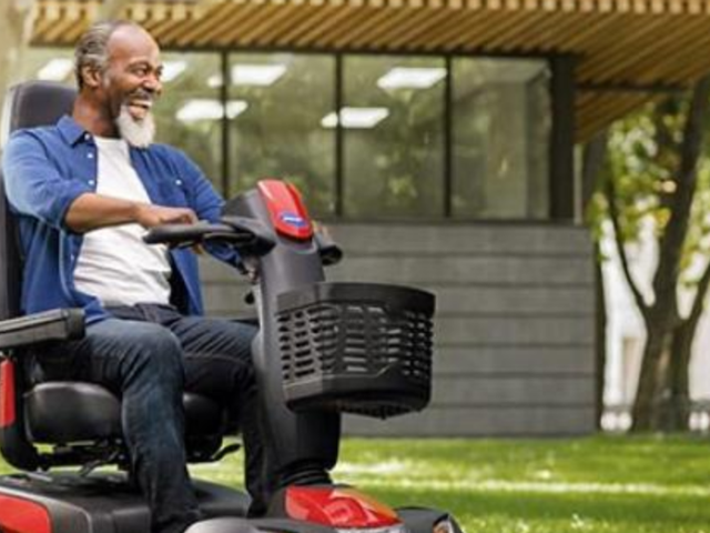 A person smiling while riding a mobility scooter in a park, with trees and a modern building in the background.