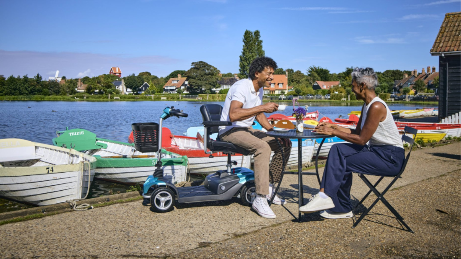 Two people enjoying a meal at a table by a scenic lake, with colorful boats moored nearby under a clear blue sky.