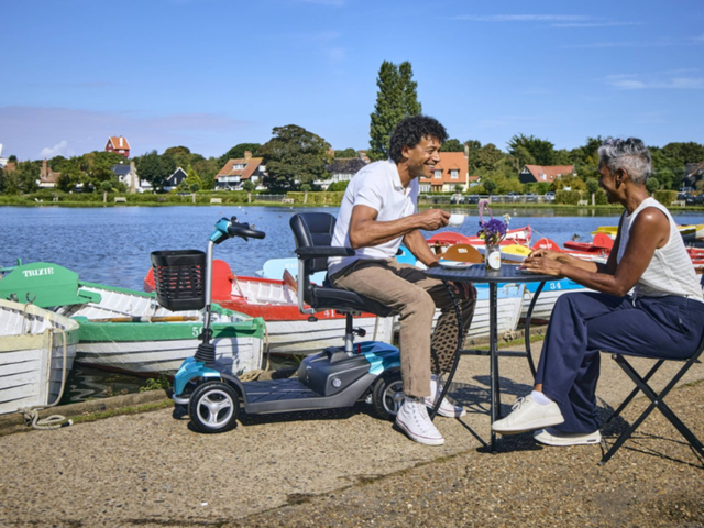 Two people enjoying a meal at a table by a scenic lake, with colorful boats moored nearby under a clear blue sky.