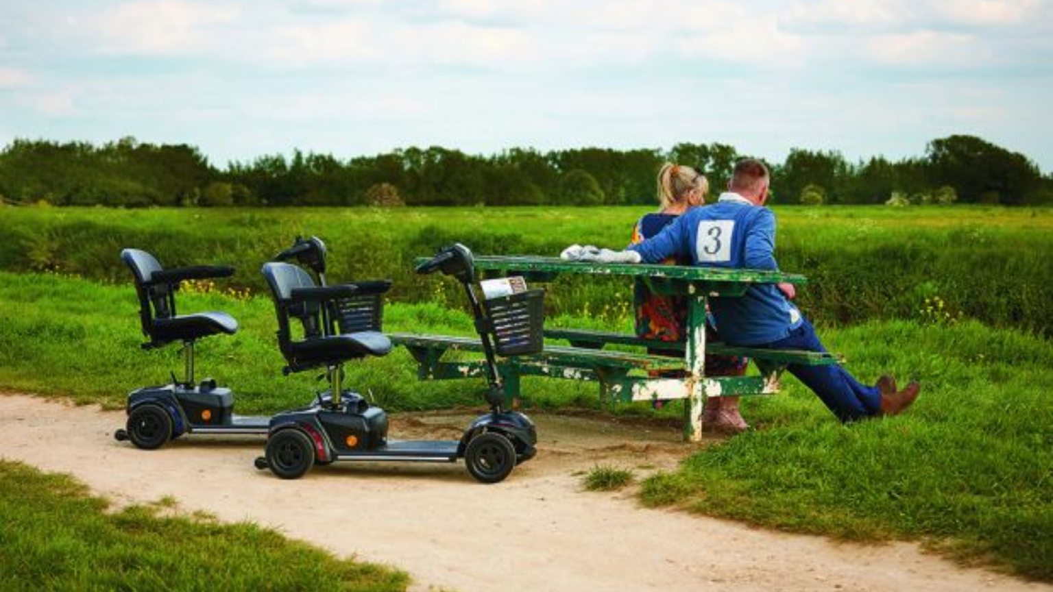 A couple sits on a green bench in a field, with two mobility scooters parked nearby.