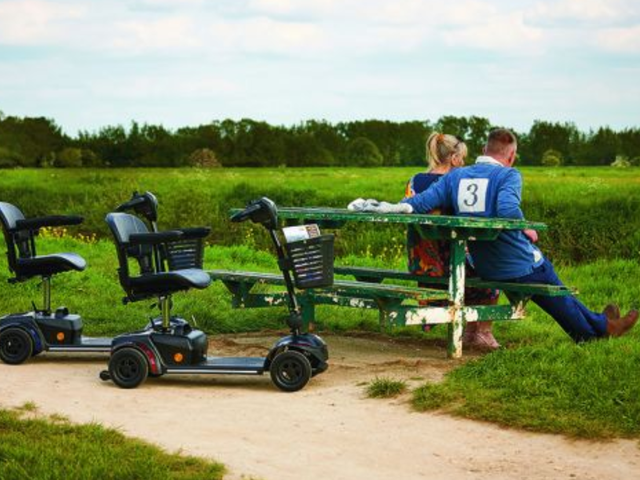 A couple sits on a green bench in a field, with two mobility scooters parked nearby.