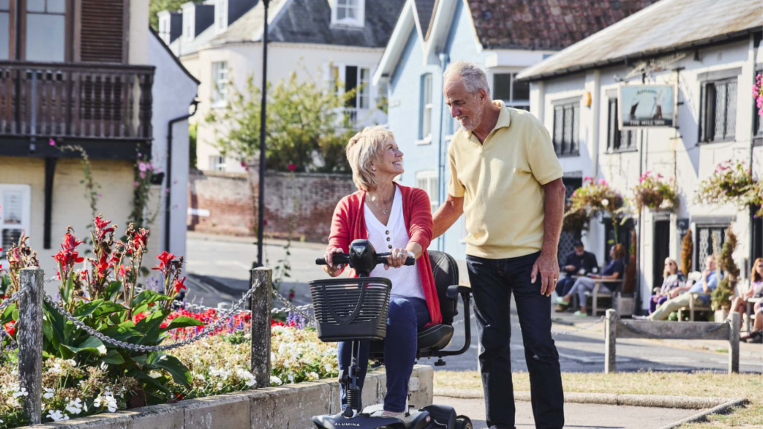 Elderly couple enjoy a sunny day; woman on mobility scooter, man walking beside her, surrounded by flowers and quaint buildings.