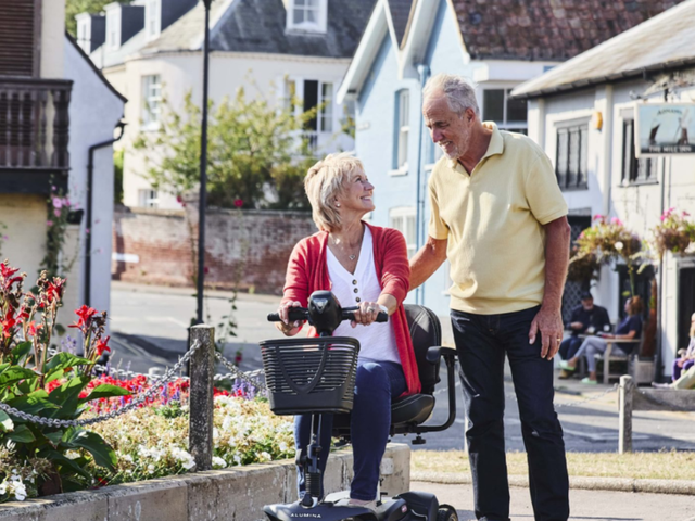 Elderly couple enjoy a sunny day; woman on mobility scooter, man walking beside her, surrounded by flowers and quaint buildings.