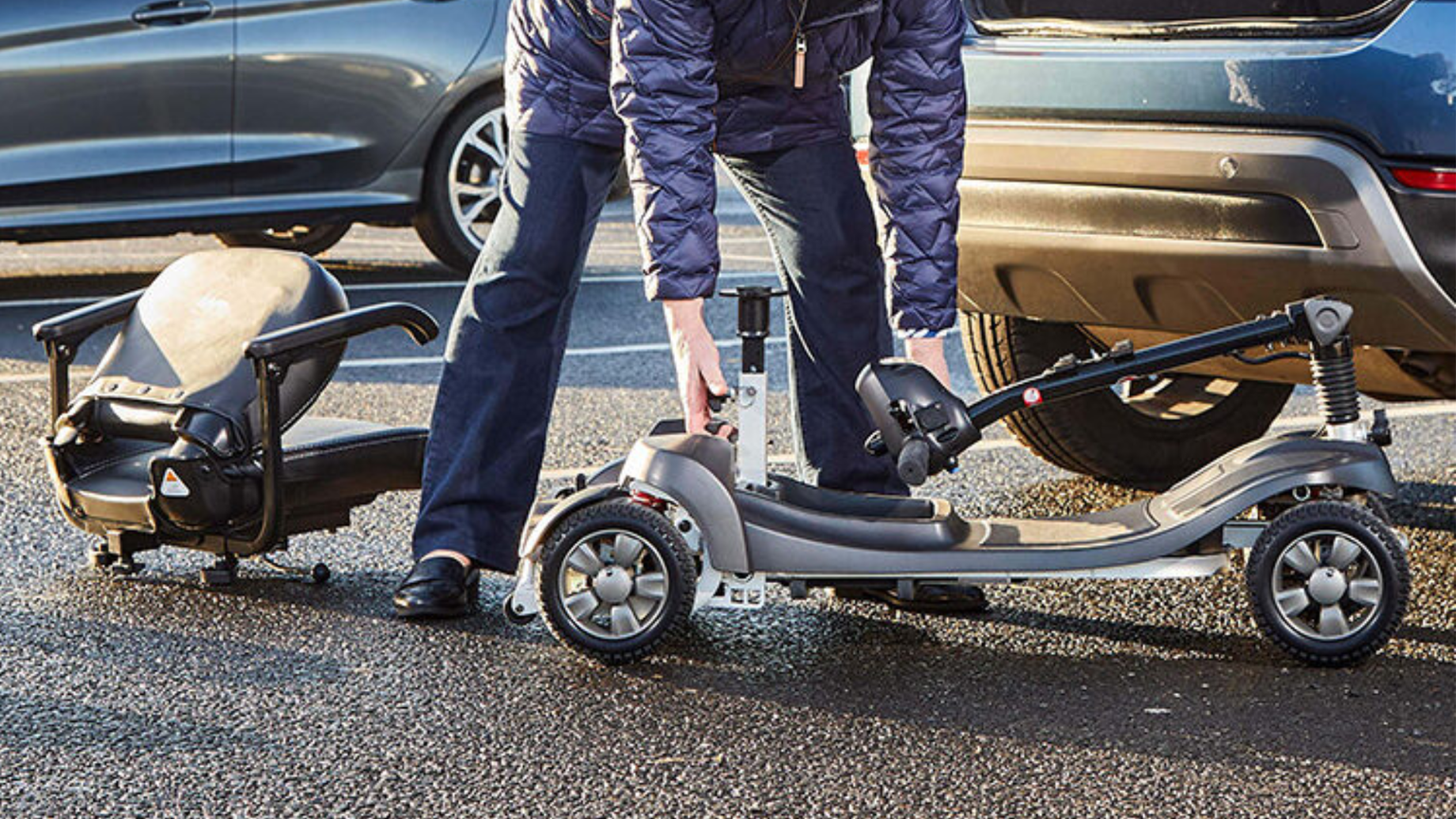 Person assembling a portable mobility scooter beside a parked car in a parking lot.