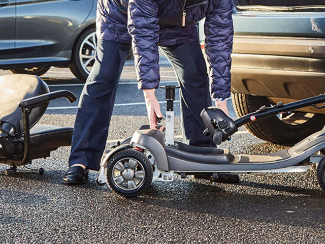 Person assembling a portable mobility scooter beside a parked car in a parking lot.