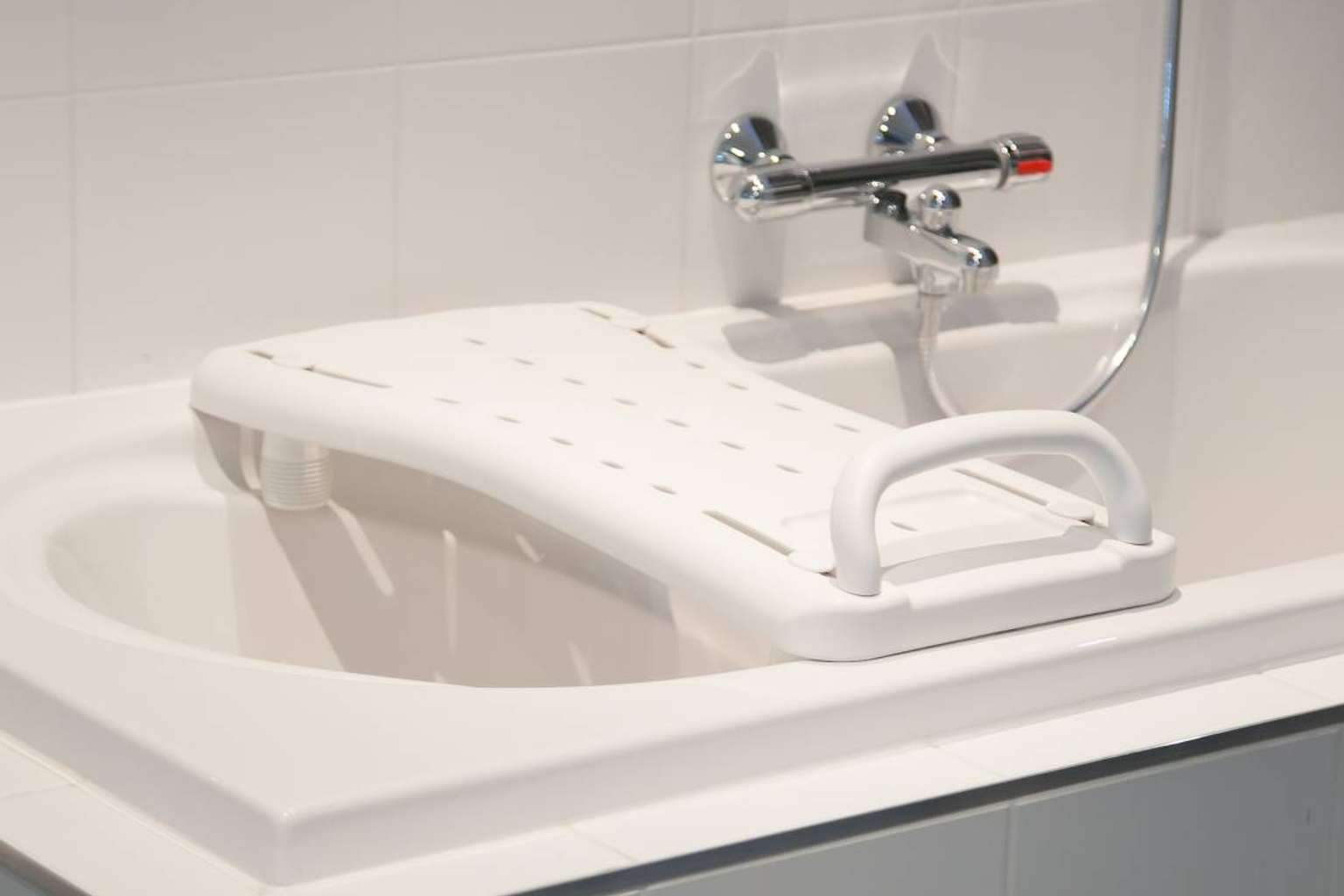 White bath board with handle placed across a bathtub, underneath a chrome faucet in a tiled bathroom.