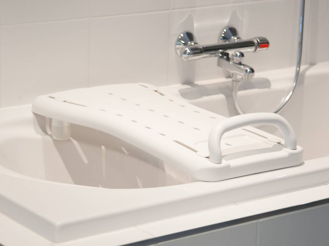 White bath board with handle placed across a bathtub, underneath a chrome faucet in a tiled bathroom.