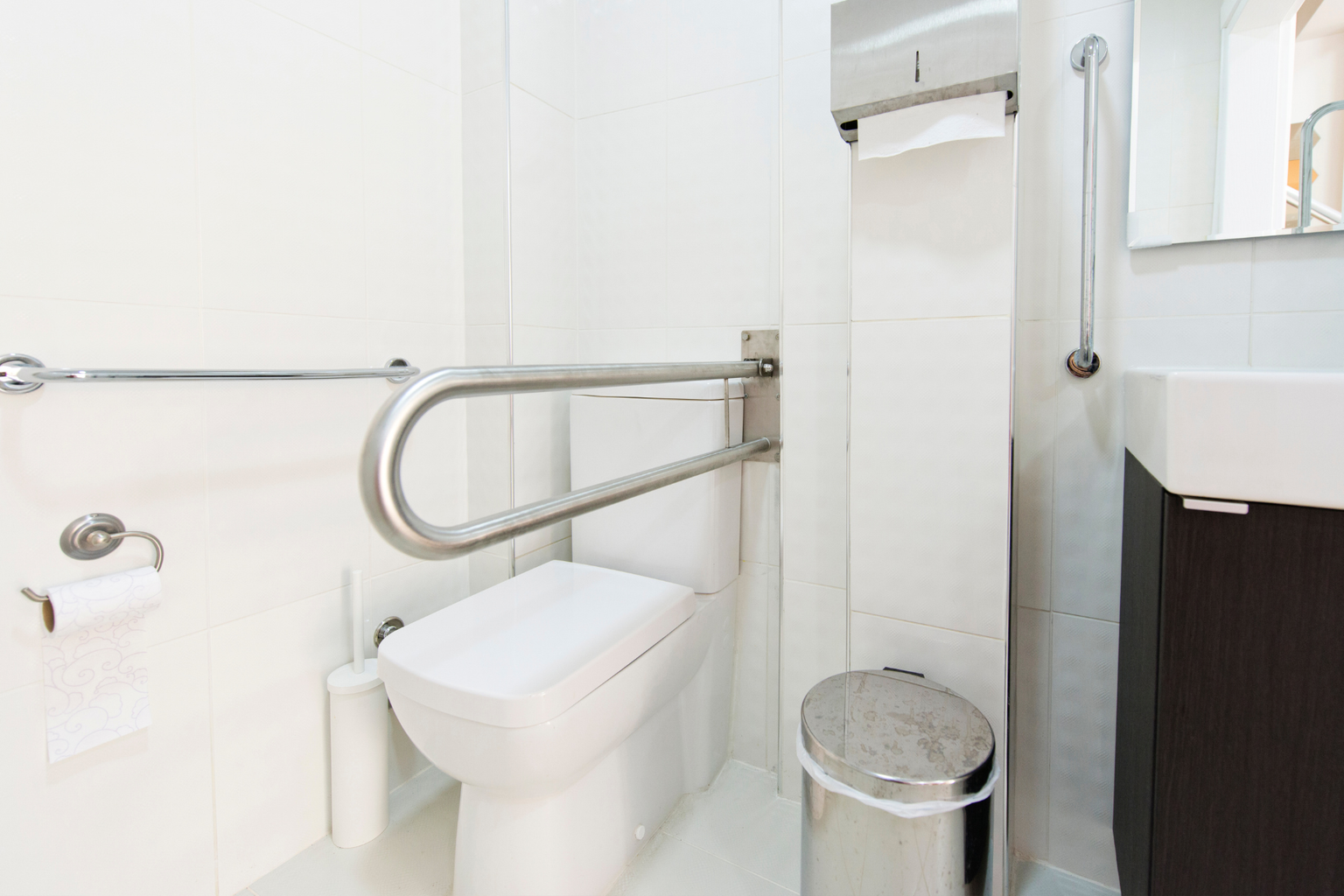 A clean, accessible bathroom with a toilet, grab bars, a trash can, toilet paper, and a sink, suitable for individuals with mobility needs.