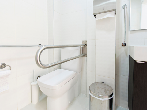 A clean, accessible bathroom with a toilet, grab bars, a trash can, toilet paper, and a sink, suitable for individuals with mobility needs.