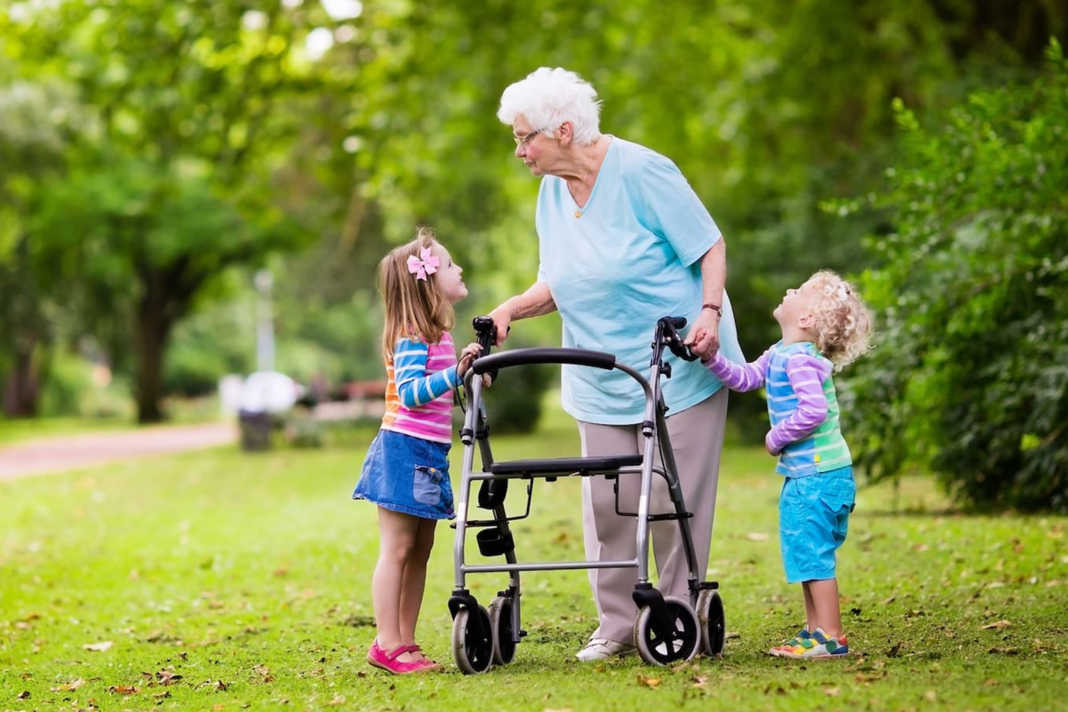 Elderly woman with a walker interacts with two young children in a park, surrounded by greenery.