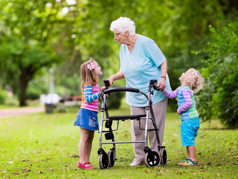 Elderly woman with a walker interacts with two young children in a park, surrounded by greenery.