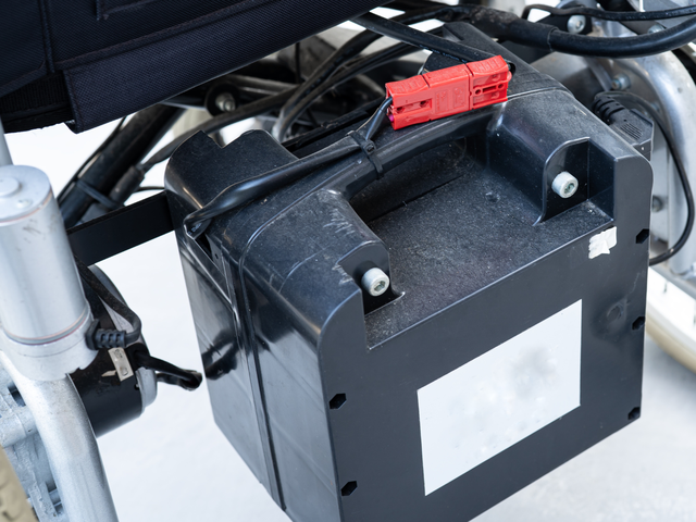 A close-up of a wheelchair battery pack attached to the wheelchair frame with visible cables and connectors.