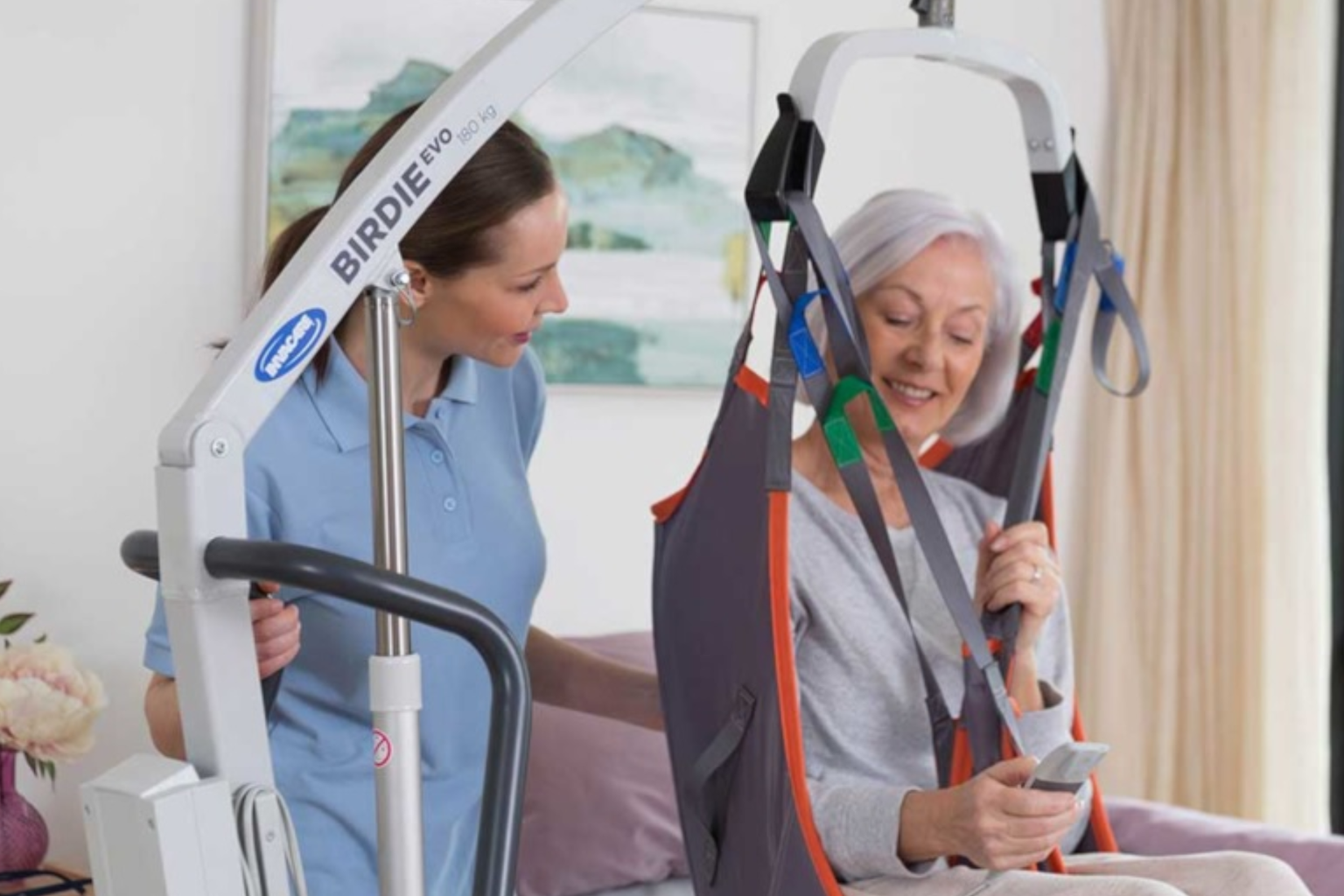 A woman using a patient lift is assisted by a caregiver in a brightly lit room. The woman is sitting comfortably in the lift, smiling.