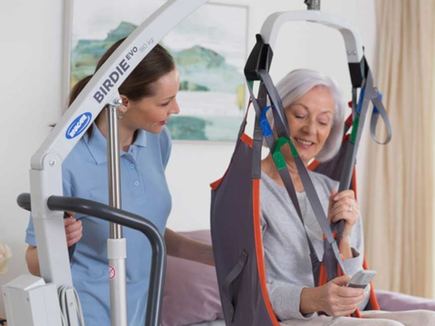 A woman using a patient lift is assisted by a caregiver in a brightly lit room. The woman is sitting comfortably in the lift, smiling.