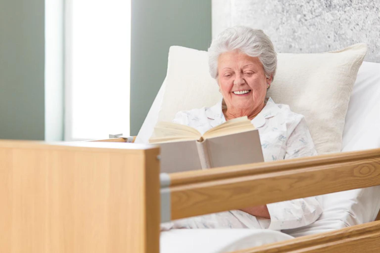 An elderly woman with gray hair smiles while reading a book in a hospital bed, wearing a light-colored pajama top.