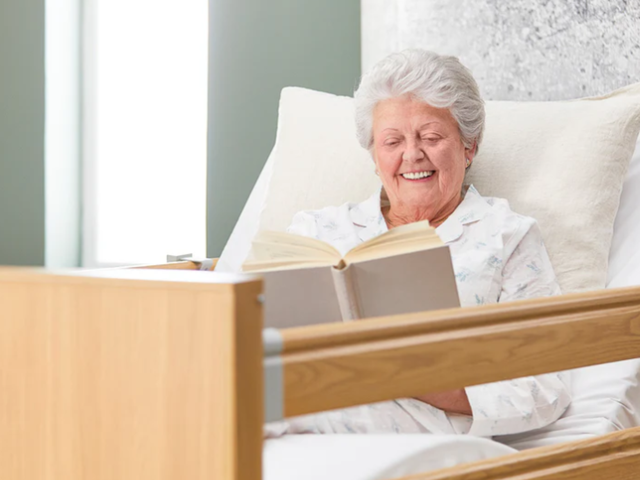 An elderly woman with gray hair smiles while reading a book in a hospital bed, wearing a light-colored pajama top.
