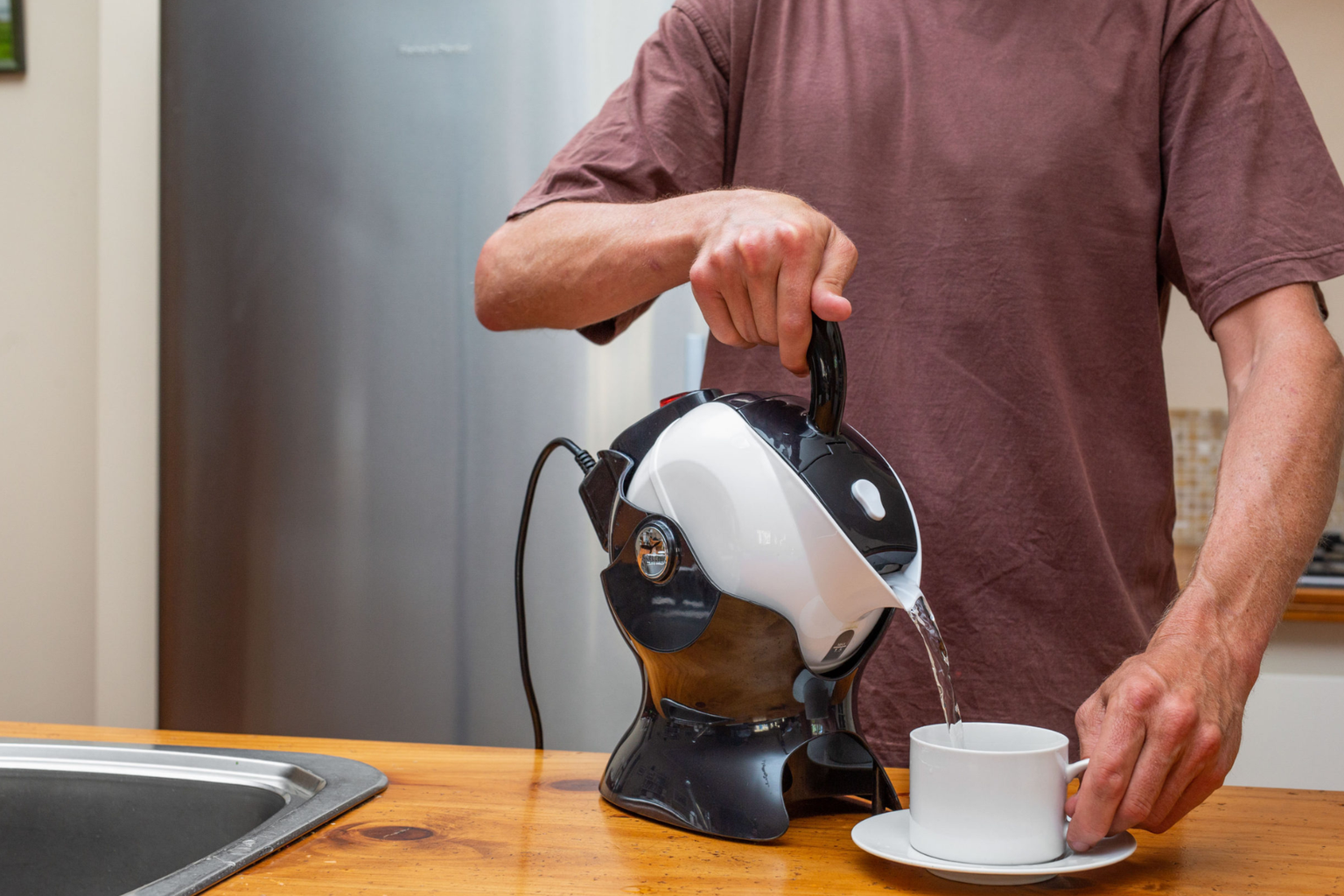 A person in a brown shirt pours hot water from a black and white kettle into a white teacup on a kitchen counter.