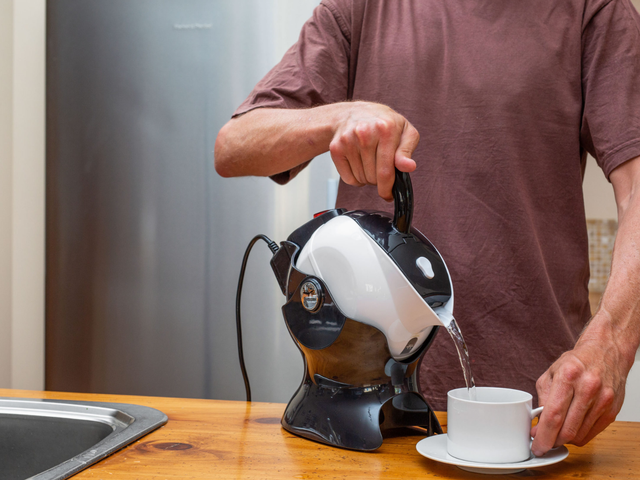 A person in a brown shirt pours hot water from a black and white kettle into a white teacup on a kitchen counter.