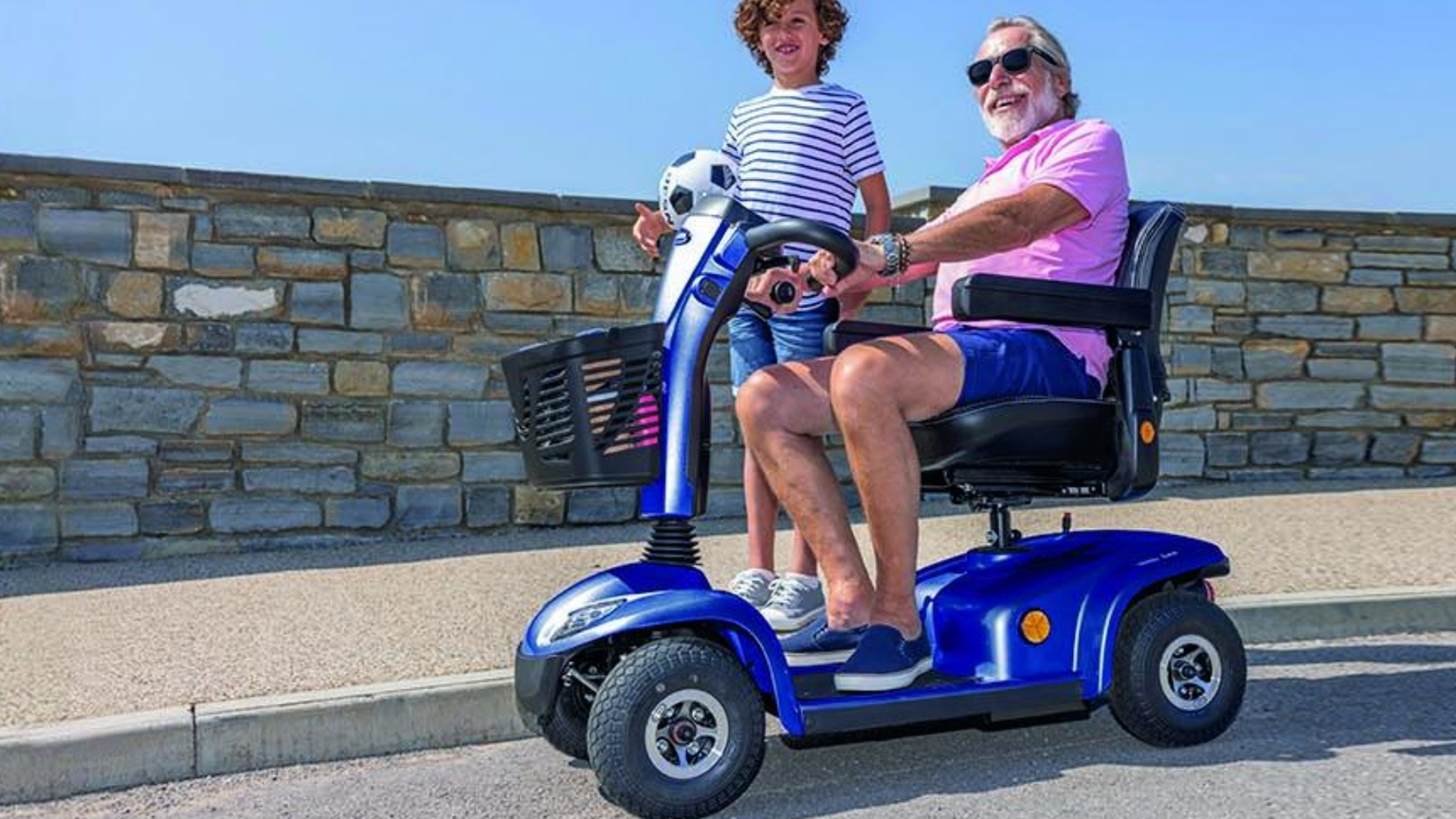 An elderly man in sunglasses rides a blue mobility scooter with a child holding a soccer ball standing beside him on a road by a stone wall.