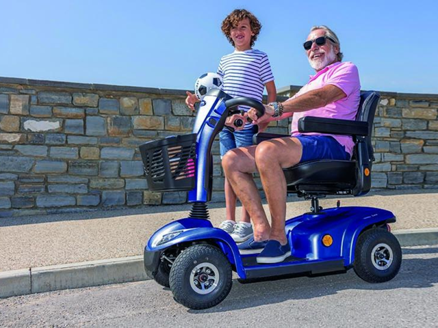 An elderly man in sunglasses rides a blue mobility scooter with a child holding a soccer ball standing beside him on a road by a stone wall.