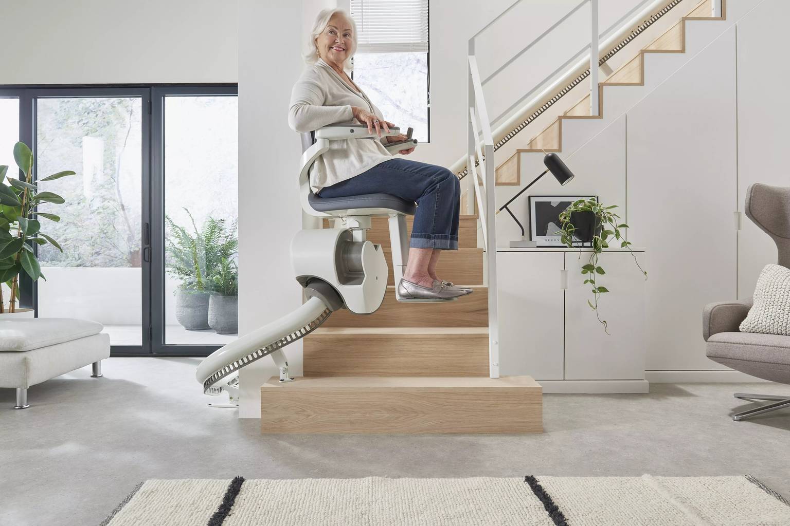An elderly woman using a stairlift, smiling in a modern, bright living room with plants and large windows.