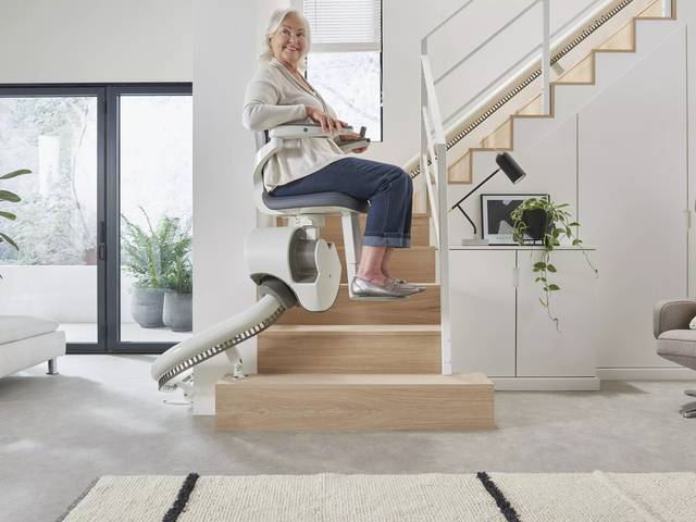 An elderly woman using a stairlift, smiling in a modern, bright living room with plants and large windows.