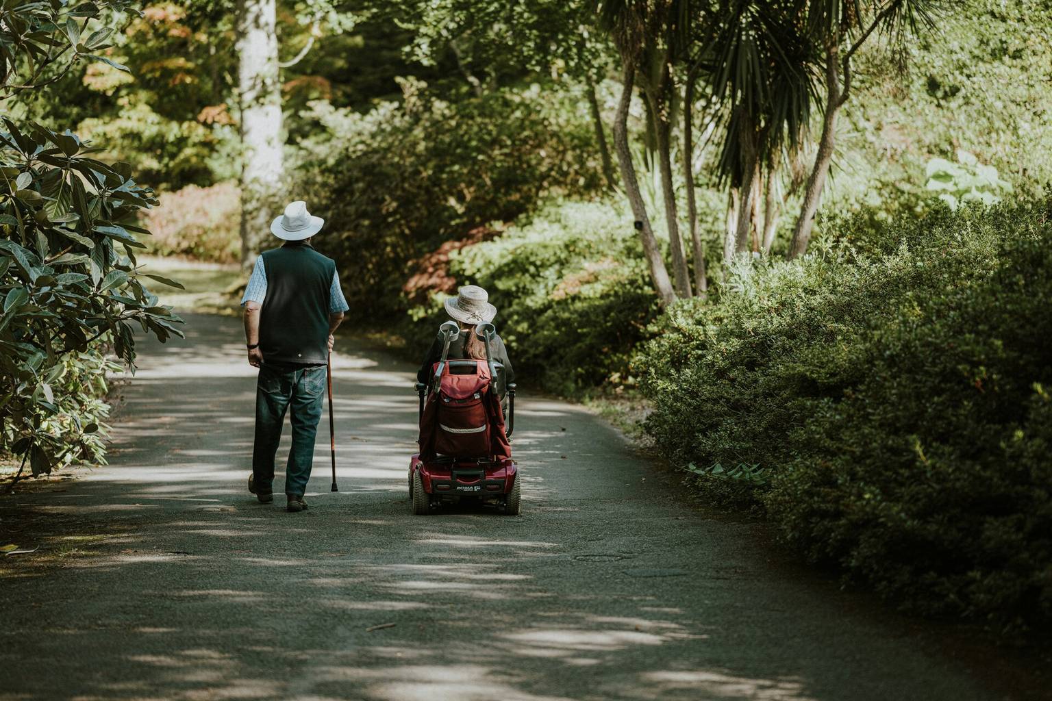Two people wearing hats, one walking with a cane and the other in a mobility scooter, strolling down a tree-lined path.
