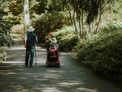 Two people wearing hats, one walking with a cane and the other in a mobility scooter, strolling down a tree-lined path.