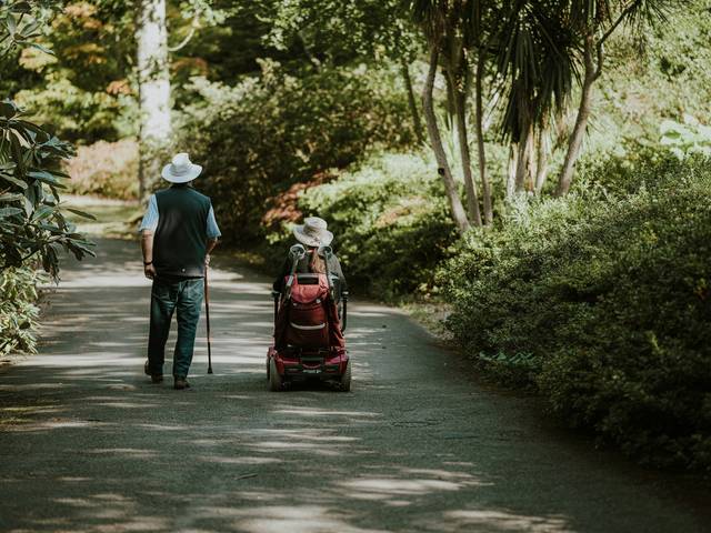 Two people wearing hats, one walking with a cane and the other in a mobility scooter, strolling down a tree-lined path.
