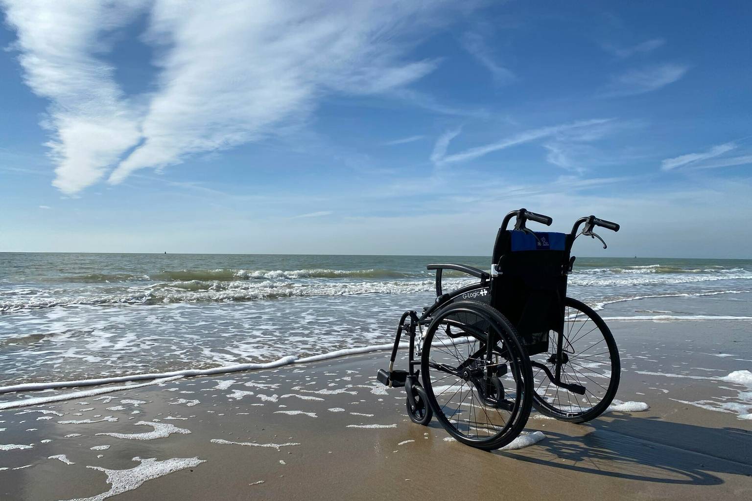 Empty wheelchair on a sandy beach near gentle waves, under a blue sky with wispy clouds.