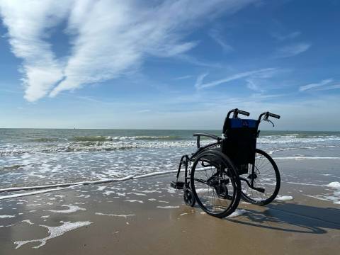 Empty wheelchair on a sandy beach near gentle waves, under a blue sky with wispy clouds.